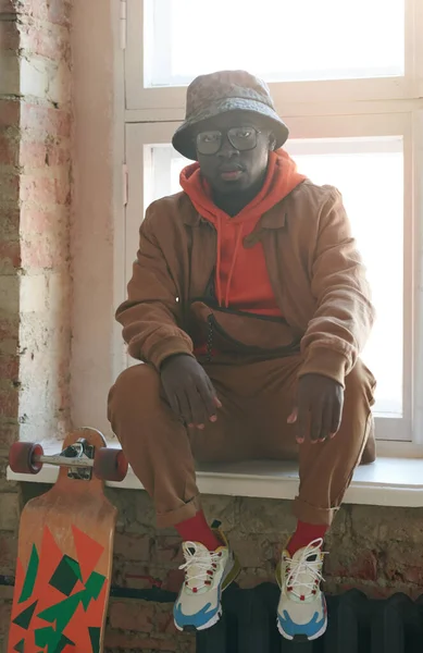 stock image Vertical medium long portrait of stylish young African American manwearing casual outfit sitting on window sill in loft room looking at camera