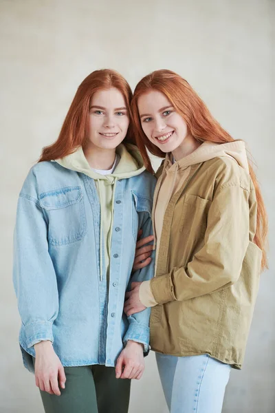 Vertical medium long portrait of two beautiful young Caucasain twin sisters with long red hair wearing casual outfits smiling at camera