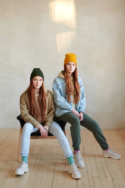 Vertical long shot portrait of young twin sisters with long red hair wearing casual outfits sitting on chair in loft studio looking at camera