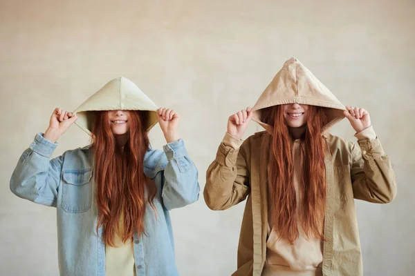 Horizontal medium srudio portrait of playful young twin sisters with long red hair hiding their eyes under hoods, copy space
