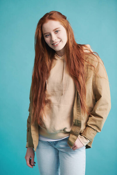 Vertical medium studio portrait of joyful young woman with long red hair wearing casual clothes posing on camera against blue wall background