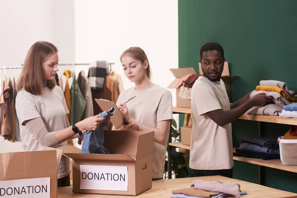 Group of three modern young volunteers working together in charity organization sorting out clothes