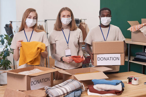 Group of young man and two women wearing protective masks working together in clothes charity organization standing at table looking at camera