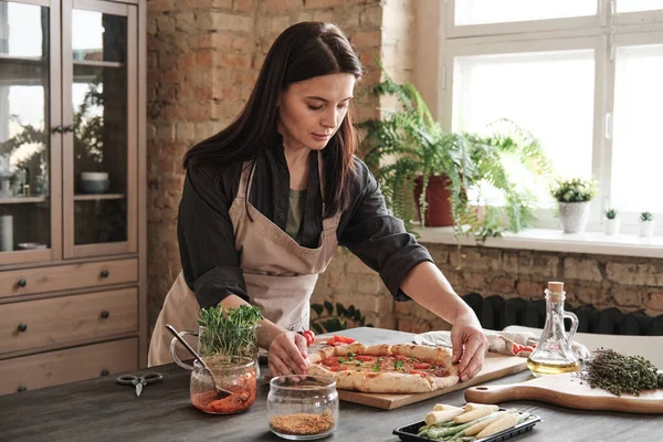 stock image Attractive brunette woman in apron standing at kitchen counter and adjusting pizza on board while preparing it for dinner