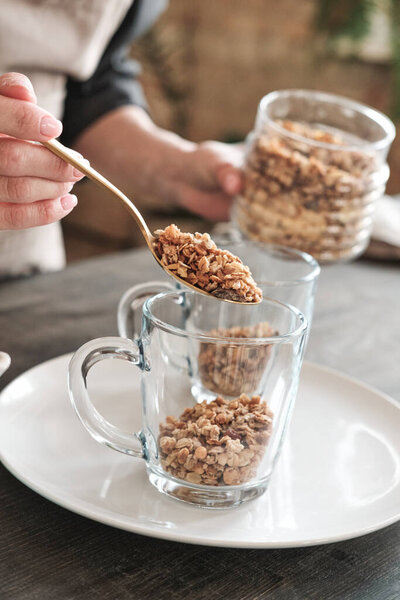 Крупный план неузнаваемой женщины, использующей ложку, когда она кладет мюсли в кружку на тарелку на кухне
