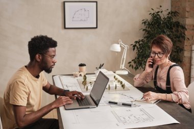 Masada oturan ve mimarlık şirketinin modern çalışma alanındaki cihazlarla çalışan çok ırklı çalışanlar.