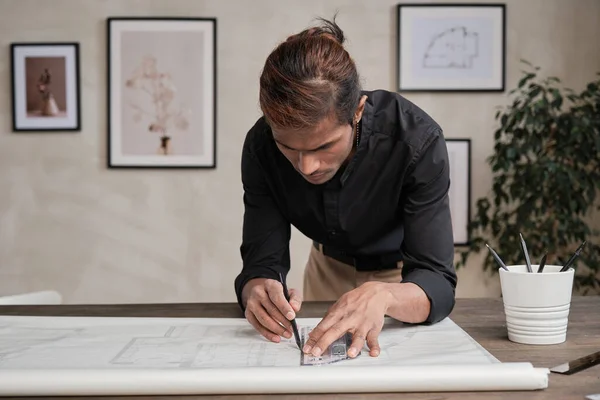 Busy young mixed race engineer in black shirt standing at table and leaning on it while using ruler and drawing line on floor plan