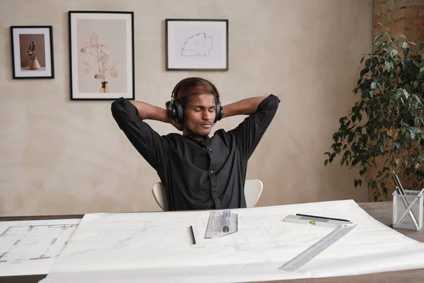 Stock image Calm young mixed race architect in wireless headphones sitting with hands behind head and taking break while working with blueprint