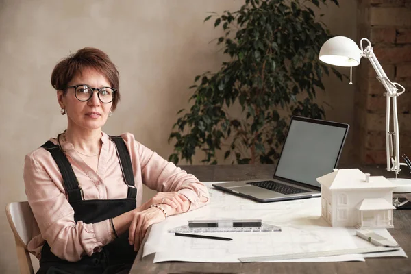 Portrait of content successful Caucasian female architect in eyeglasses sitting at table with laptop, D model of house and blueprint in office
