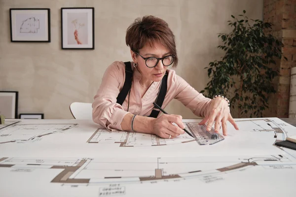Serious mature woman with short hair sitting at table in loft office and using ruler while making blueprint drawing