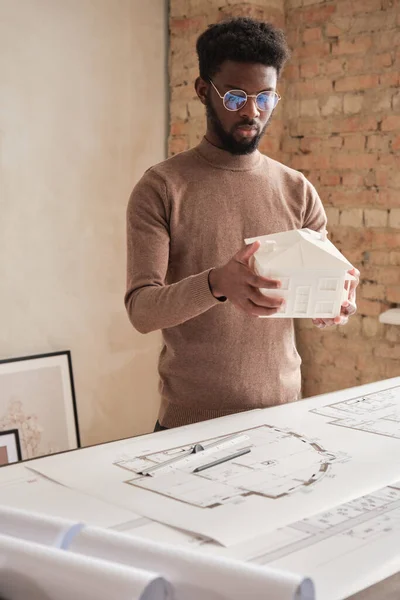 Serious creative young African-American building designer with beard standing at table with floor plan and looking at D model of house