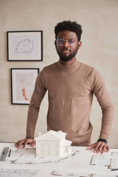 Portrait of content young African-American building architect in glasses standing at table with floor plan and D model of house