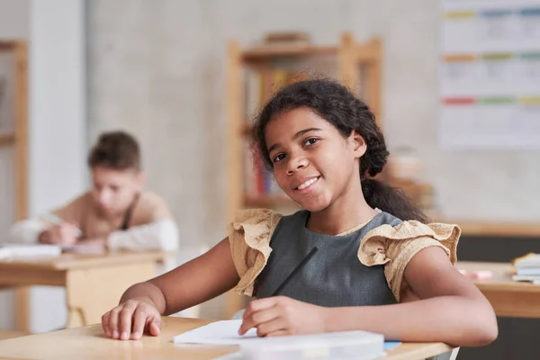 stock image Portrait of smiling mixed race girl looking at camera while sitting at desk in classroom with wooden decor, copy space