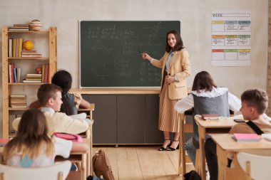 Sınıfta tahtayı işaret eden ve bir grup çocukla konuşan gülümseyen bayan öğretmenin tam boy portresi.