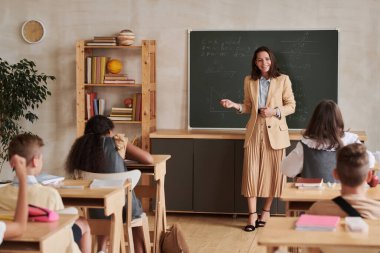 Genç bayan öğretmenin tam boy portresi tahtada duruyor ve sınıftaki çocuklarla konuşuyor, boşluğu kopyala