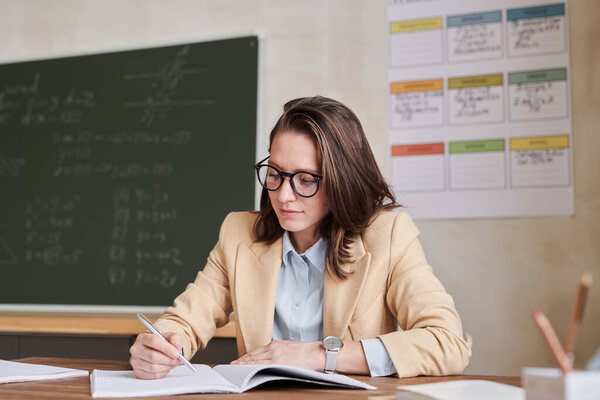 Warm-toned portrait of young female teacher sitting at desk in school classroom and writing, copy space