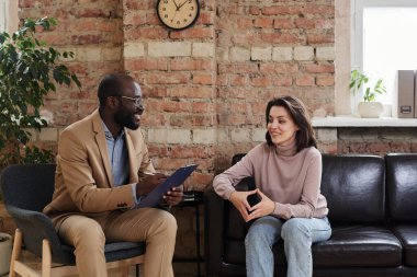 Gülümseyen genç bir kadın koltukta oturup parmaklarını birbirine değdirirken psikologla duyguları tartışıyor.
