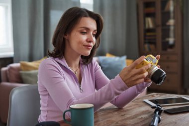 Gülümseyen beyaz kadın oturma odasında oturuyor ve fotoğraf çekiminden sonra dijital kamerada resimlere bakıyor.