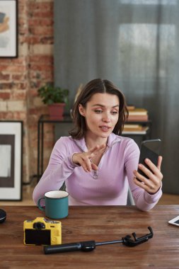 Kendine güvenen genç bayan fotoğrafçı ahşap masada oturuyor ve akıllı telefondaki video bağlantısıyla çevrimiçi danışmanlık yaparken el kol hareketi yapıyor.