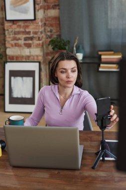 Kahverengi saçlı, güzel, beyaz bir kadın dizüstü bilgisayarla masada oturuyor ve canlı yayında yorumlar okuyor.