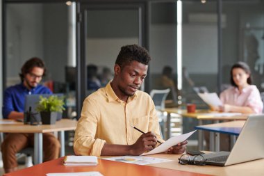 Meşgul genç Afro-Amerikalı iş uzmanı masada oturuyor ve açık alan ofisindeki evrakları inceliyor.
