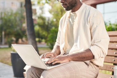 Sakallı Afrikalı-Amerikalı SMM yöneticisi kentteki bankta oturuyor ve dizüstü bilgisayarında yazıyor.