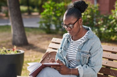 Gözlüklü ciddi bir Afro-Amerikalı öğrenci. Şehir parkında bankta oturuyor ve günlük tutuyor.