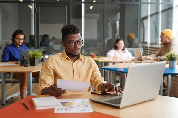 Gözlüklü genç siyah analist masada oturuyor ve birlikte çalışma alanındaki kağıtları incelerken dizüstü bilgisayar kullanıyor.