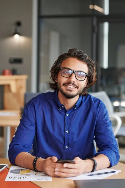 stock image Portrait of cheerful young mixed race businessman with beard wearing blue shirt using smartphone at table in office