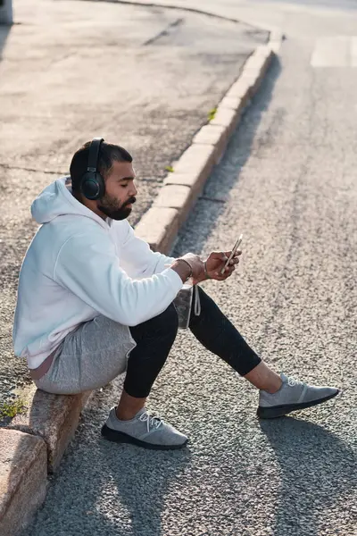Calm young bearded man in white hoodie sitting on curb and using smartphone while enjoying music in headphones