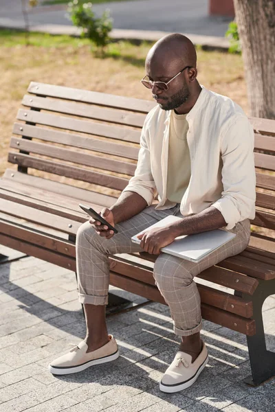 严肃的非洲裔美国企业家穿着时髦的休闲装坐在城市的长椅上 用智能手机工作 — 图库照片