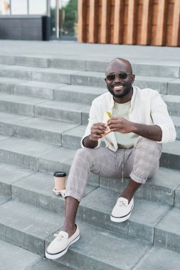 Güneş gözlüklü gülümseyen siyah bir adamın portresi kahve fincanı ile merdivenlerde oturuyor ve sandviç yiyor.