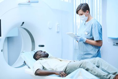 Önlüklü ve maskeli beyaz hemşire MRI tarayıcısında duruyor ve Afro-Amerikalı hastaya belirtileri soruyor.