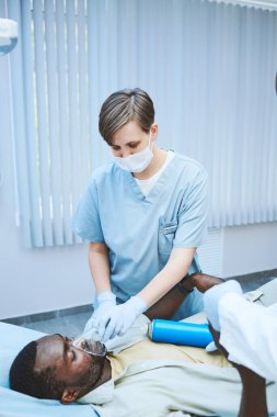 Cerrahi maskeli ve eldivenli genç hemşire hastanede oksijen seviyesini arttırmak için siyah hastaya oksijen maskesi veriyor.