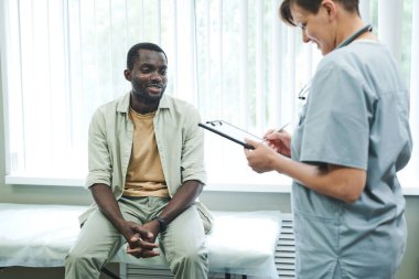 Önlüklü gülümseyen genç hemşire tıbbi kayıtları doldururken not alıyor ve hastalarla konuşuyor.