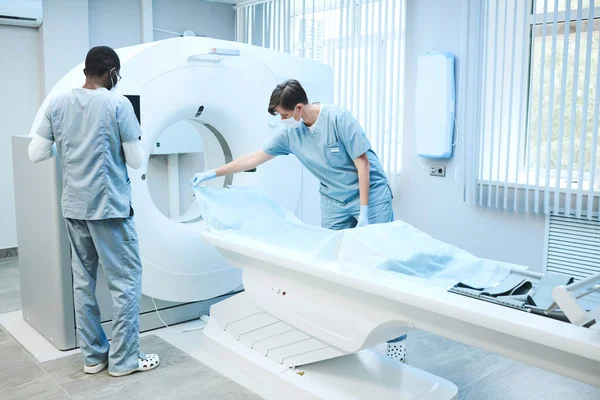 Maskeli ve önlüklü çok ırklı hemşireler hastanın muayenesi için MRI taraması hazırlıyorlar: Masaya tek kullanımlık çarşaf seren kadın.