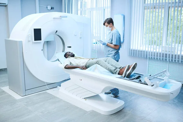 MRI specialist in facial mask and gloves standing at patient lying on table and filling his card online