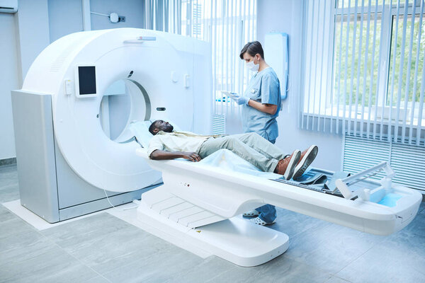 Специалист по магнитно-резонансной томографии в маске и перчатках, стоящий у лежащего на столе пациента и заполняющий его карту онлайн