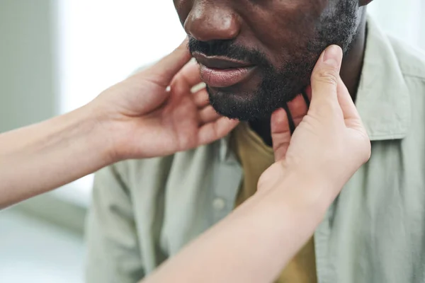 Close-up of unrecognizable nurse palpating chin of black man with stubble at medical checkup
