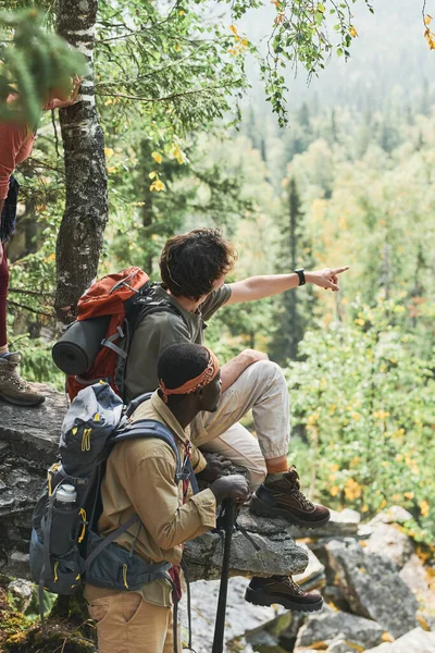背负背包的年轻远足者坐在石头上 向黑人朋友展示森林风景的背景图 — 图库照片
