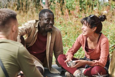 Bir grup neşeli genç ırklar arası yürüyüşçü yerde oturup yemek yerken sohbet ediyorlar.