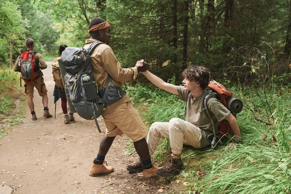 徒步旅行的非裔美国人 背着背包 在帮助倒下的男子站起来进行森林远足时 向他伸出了援助之手 — 图库照片