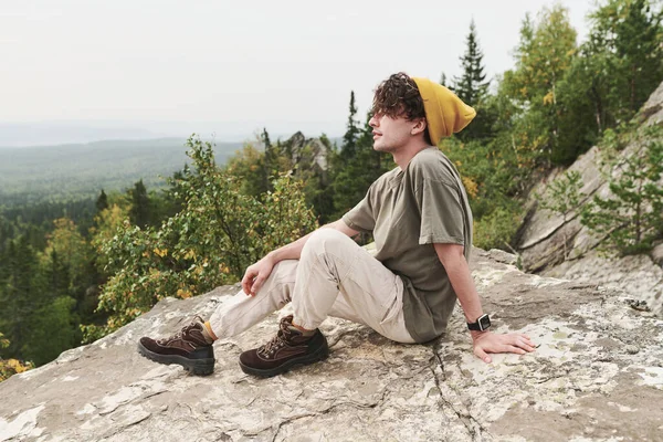 白种人年轻的梦游者 头戴黄帽 脚踏黄靴 坐在大石头上 凝视着森林风光 — 图库照片