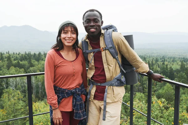 快乐的多种族夫妇站在栏杆边与森林景观相映成趣 欣赏着远足的相机 — 图库照片