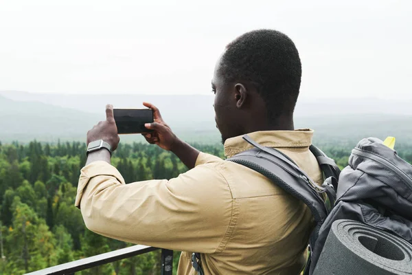 年轻的非洲裔美国人背着背包站在桥上 用电话拍摄森林景观 — 图库照片