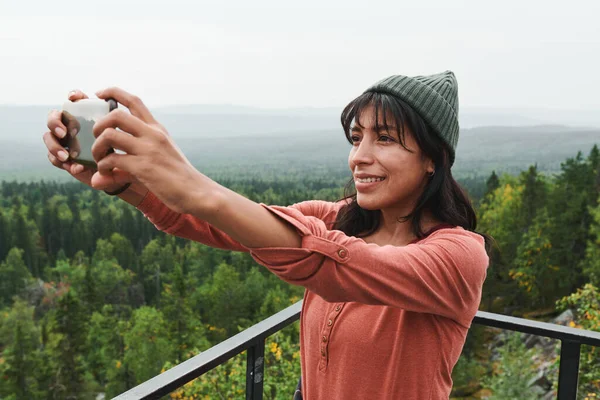 戴着礼帽站在桥上微笑着年轻的混血女子 并在户外用智能手机拍照 — 图库照片