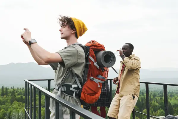 年轻的徒步旅行者背着背包站在桥上微笑 一边在电话上拍摄风景 一边拯救生命 — 图库照片