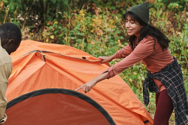 正面的年轻混血女子戴着帽子指着杆子 同时帮助男友在露营时搭起帐篷 — 图库照片
