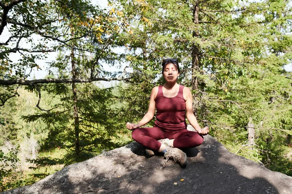 Serene young woman with closed eyes sitting with crossed legs and holding hands in mudra while meditating in mountains