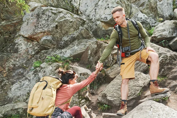 两对年轻夫妇背着背包一起爬上岩石山 男人牵着女友的手帮助她爬上石头 — 图库照片